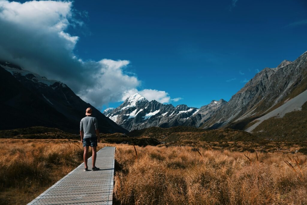 Person walks on wooden path with Mount Cook in the background under a blue sky.